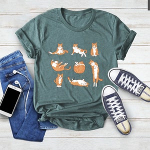 May include: Teal green t-shirt featuring a playful design of orange and white cats in various poses. The shirt is paired with blue jeans, white wired earbuds, and navy blue canvas sneakers.