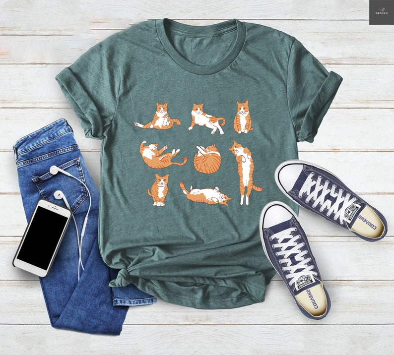 May include: Teal green t-shirt featuring a playful design of orange and white cats in various poses. The shirt is paired with blue jeans, white wired earbuds, and navy blue canvas sneakers.