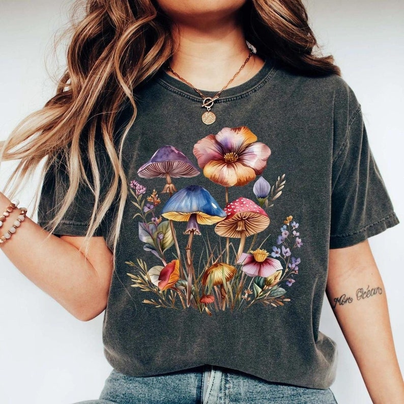 May include: Dark gray t-shirt featuring a colorful illustration of mushrooms and flowers. The design includes purple, blue, red, and orange mushrooms, with a variety of flowers and foliage. The shirt has a vintage, worn look.