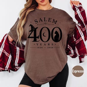 Puede incluir: Una camiseta de manga corta color espresso con un estampado gr&aacute;fico negro que dice SALEM MASSACHUSETTS 400 YEARS 1626-2026. El dise&ntilde;o incluye un sombrero de bruja, una luna creciente y la silueta de un gato integrada en el n&uacute;mero 400.