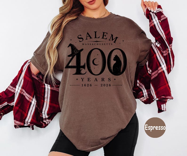 Puede incluir: Una camiseta de manga corta color espresso con un estampado gr&aacute;fico negro que dice SALEM MASSACHUSETTS 400 YEARS 1626-2026. El dise&ntilde;o incluye un sombrero de bruja, una luna creciente y la silueta de un gato integrada en el n&uacute;mero 400.