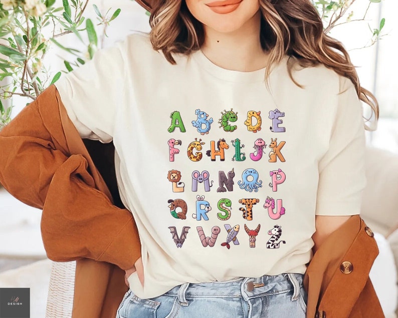 May include: White t-shirt with a colorful alphabet design featuring animals. Each letter is formed by a different animal, such as a giraffe, a penguin, a worm, and a zebra.