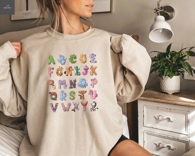 May include: Beige sweatshirt featuring a colorful alphabet design with each letter represented by a different animal. The letters are in a playful font, making it a fun and educational garment. The sweatshirt is a comfortable, casual style.