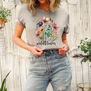 May include: Athletic heather gray t-shirt featuring a watercolor floral design with the word "wildflower" in a script font. The flowers are in shades of pink, purple, and blue. The shirt has rolled-up sleeves.