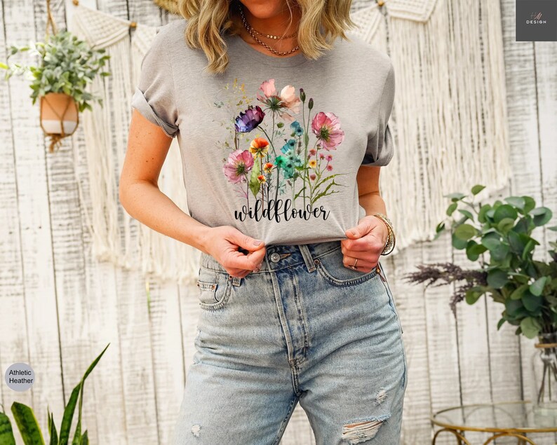 May include: Athletic heather gray t-shirt featuring a watercolor floral design with the word "wildflower" in a script font. The flowers are in shades of pink, purple, and blue. The shirt has rolled-up sleeves.