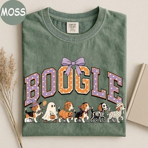 K&ouml;nnte beinhalten: Ein moosgr&uuml;nes T-Shirt mit dem Schriftzug BOOGLE in orangefarbenen und violetten gepunkteten Buchstaben und einer violetten Schleife dar&uuml;ber. Darunter sind sechs Beagle-Hunde in Halloween-Kost&uuml;men abgebildet: Hexe, Geist, K&uuml;rbis, Vampir, Skelett und Mumie.
