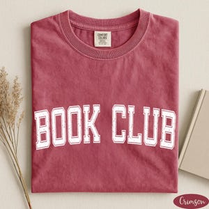 May include: A crimson short-sleeved t-shirt with a crew neckline featuring the white text BOOK CLUB printed in a collegiate font across the chest. The shirt has a Comfort Colors brand label on the inside collar and a weathered, textured fabric finish.