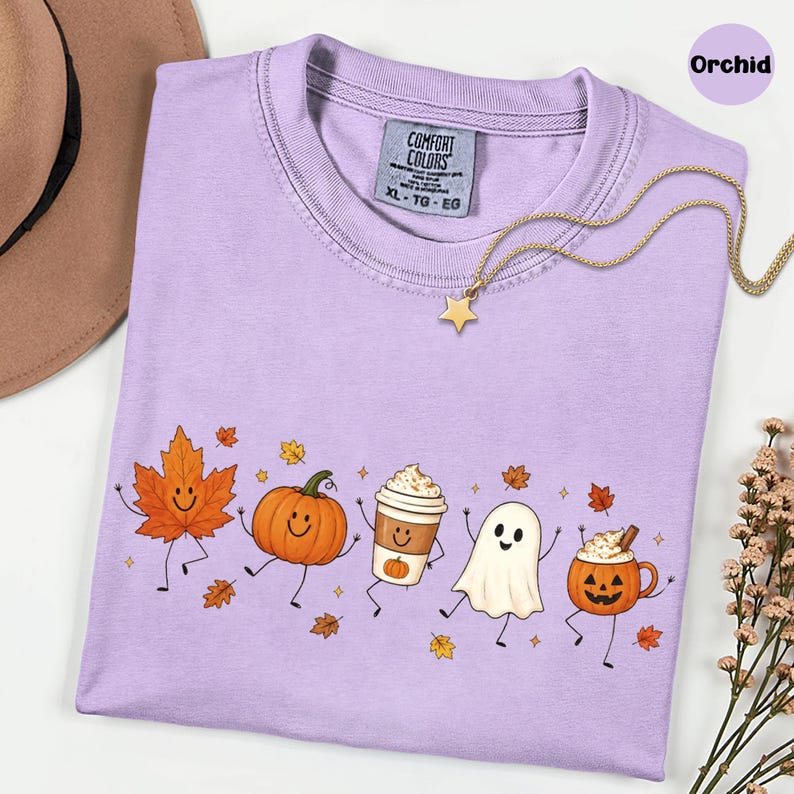 K&ouml;nnte beinhalten: Ein hellviolettes Kurzarm-T-Shirt mit einem Grafikdruck, der ein l&auml;chelndes Ahornblatt, einen K&uuml;rbis, einen Kaffeebecher, ein Gespenst und eine Jack-o'-Lantern-Tasse zeigt, die alle mit Armen und Beinen versehen sind.