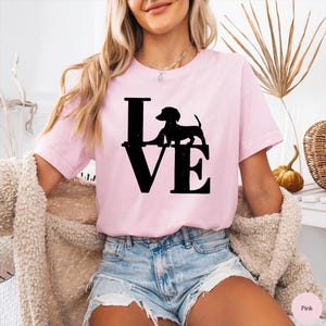 May include: A light pink t-shirt with the word "LOVE" in large black letters. A black silhouette of a dachshund dog is positioned above the "VE". The shirt is short-sleeved and made of a soft material.