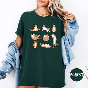 May include: Dark green t-shirt featuring a playful design of orange and white cats in various poses. The cats are illustrated in a cartoon style, adding a whimsical touch to the shirt. The word "FOREST" is printed on a green circle.