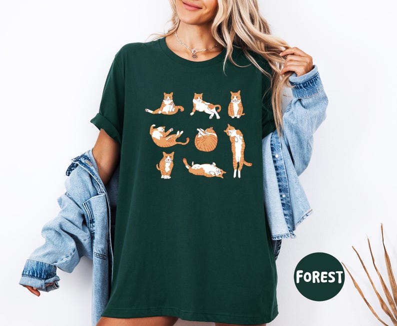 May include: Dark green t-shirt featuring a playful design of orange and white cats in various poses. The cats are illustrated in a cartoon style, adding a whimsical touch to the shirt. The word "FOREST" is printed on a green circle.