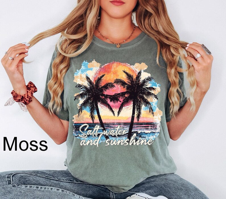 May include: Moss green t-shirt featuring a sunset scene with palm trees and the text "Saltwater and sunshine." The graphic includes a vibrant orange and yellow sun, blue water, and colorful clouds. The shirt is a relaxed fit.