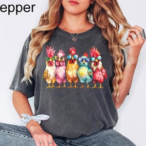 Puede incluir: Una camiseta gris oscuro con un gr&aacute;fico de cinco pollos con gafas de sol. Los pollos est&aacute;n en fila, mirando hacia adelante, y todos son de diferentes colores. El texto "Pepper" est&aacute; en la esquina superior izquierda de la imagen.