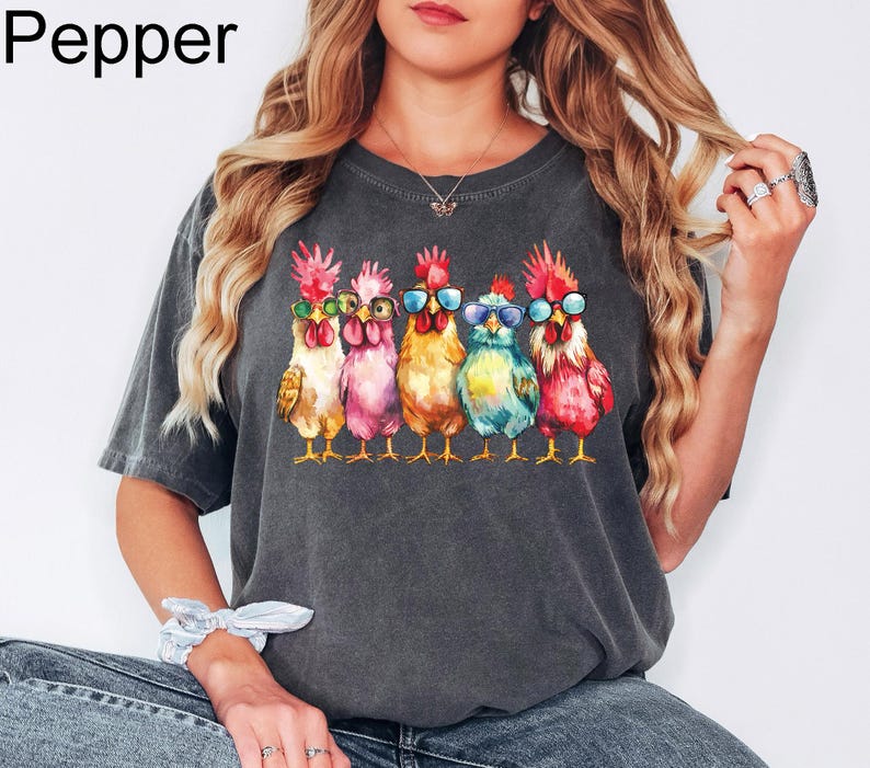 Puede incluir: Una camiseta gris oscuro con un gr&aacute;fico de cinco pollos con gafas de sol. Los pollos est&aacute;n en fila, mirando hacia adelante, y todos son de diferentes colores. El texto "Pepper" est&aacute; en la esquina superior izquierda de la imagen.