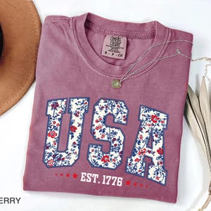 May include: Berry-colored t-shirt with the word "USA" in a floral design. The shirt includes the text "EST. 1776" and is paired with a silver necklace. A brown hat and dried plant are in the background.