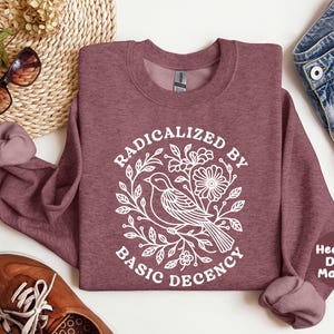 May include: Heather dark maroon sweatshirt with the text "RADICALIZED BY BASIC DECENCY" in white. The design includes a bird, flowers, and leaves. The sweatshirt is displayed with sunglasses, shoes, and denim shorts.