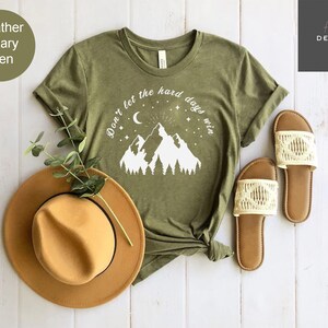 Pu&ograve; includere: T-shirt verde militare melange con grafica di montagna e la scritta "Don't let the hard days win". Un cappello fedora marrone e un paio di sandali color crema sono accanto alla maglietta. La maglietta &egrave; annodata in basso.
