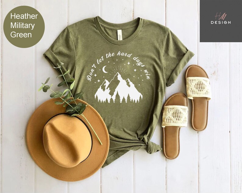 Pu&ograve; includere: T-shirt verde militare melange con grafica di montagna e la scritta "Don't let the hard days win". Un cappello fedora marrone e un paio di sandali color crema sono accanto alla maglietta. La maglietta &egrave; annodata in basso.