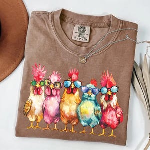 Puede incluir: Una camiseta marr&oacute;n con una ilustraci&oacute;n de acuarela de cinco pollos con gafas de sol. Los pollos est&aacute;n en fila, y cada uno tiene un cuerpo y gafas de sol de color diferente. El texto "COMFORT COLORS" est&aacute; impreso en la camiseta.