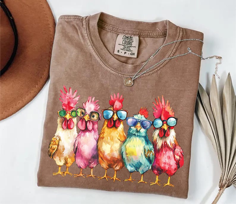 Puede incluir: Una camiseta marr&oacute;n con una ilustraci&oacute;n de acuarela de cinco pollos con gafas de sol. Los pollos est&aacute;n en fila, y cada uno tiene un cuerpo y gafas de sol de color diferente. El texto "COMFORT COLORS" est&aacute; impreso en la camiseta.