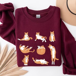 May include: A maroon long-sleeve crewneck sweatshirt featuring a graphic print of eight orange and white tabby cats in various poses. The cats are depicted sitting, stretching, rolling, and standing. The garment has a ribbed collar and cuffs.