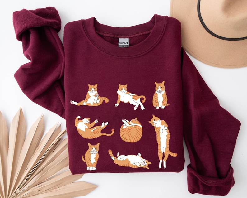 May include: A maroon long-sleeve crewneck sweatshirt featuring a graphic print of eight orange and white tabby cats in various poses. The cats are depicted sitting, stretching, rolling, and standing. The garment has a ribbed collar and cuffs.