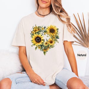 Pode incluir: Camiseta creme com um design em forma de cora&ccedil;&atilde;o composto por girass&oacute;is amarelos e folhas verdes. A palavra "Natural" est&aacute; impressa no canto inferior direito. A camisa tem mangas curtas e um corte folgado.