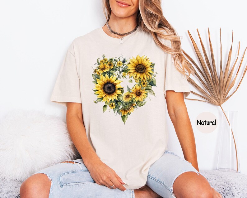 Pode incluir: Camiseta creme com um design em forma de cora&ccedil;&atilde;o composto por girass&oacute;is amarelos e folhas verdes. A palavra "Natural" est&aacute; impressa no canto inferior direito. A camisa tem mangas curtas e um corte folgado.