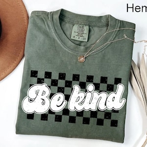 May include: A green t-shirt with a black and white checkered pattern and the words "Be kind" in a retro white font.