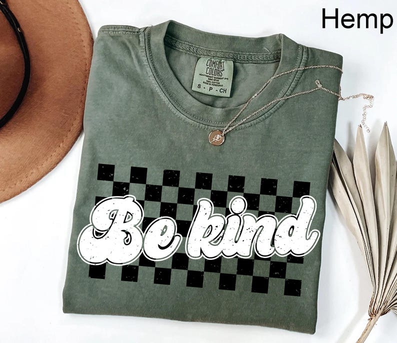 Pu&ograve; includere: Una t-shirt a maniche corte color canapa con una grafica a scacchi bianchi e neri e la scritta "Be kind" in un carattere bianco, usurato e arrotondato. La maglietta presenta un girocollo e un'etichetta del marchio Comfort Colors sul colletto.