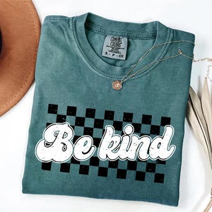 Pu&ograve; includere: Una t-shirt a maniche corte color verde acqua con scollo rotondo ed etichetta Comfort Colors. La parte anteriore presenta una grafica a scacchi nera e verde acqua con la scritta bianca "Be kind" in un carattere arrotondato.