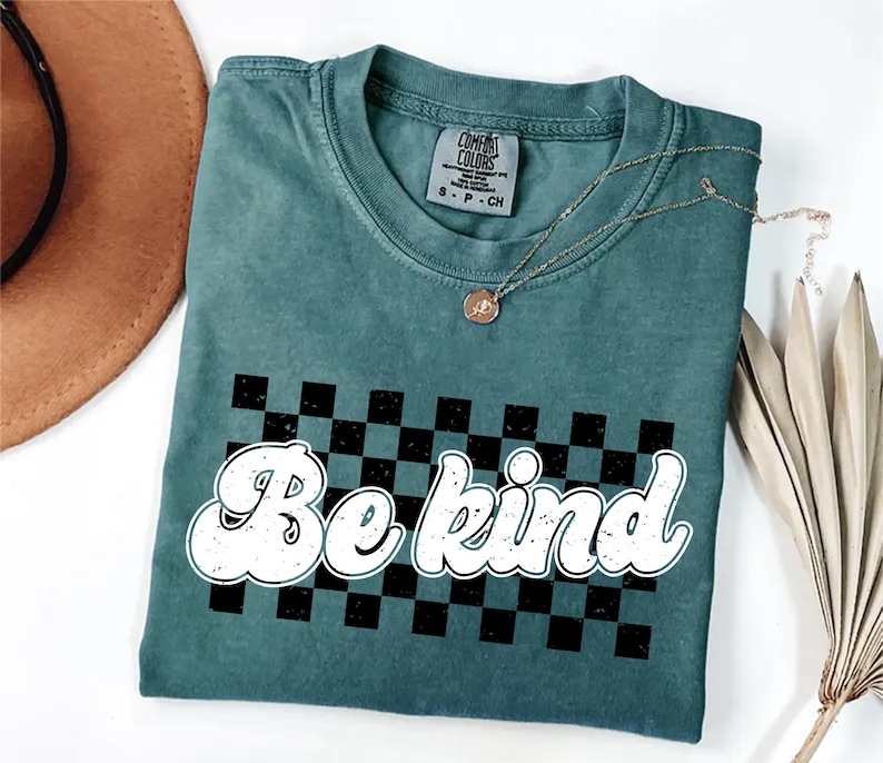 Pu&ograve; includere: Una t-shirt a maniche corte color verde acqua con scollo rotondo ed etichetta Comfort Colors. La parte anteriore presenta una grafica a scacchi nera e verde acqua con la scritta bianca "Be kind" in un carattere arrotondato.