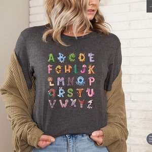 May include: Dark gray t-shirt featuring a colorful alphabet design. Each letter is creatively illustrated as an animal or creature, including a giraffe, octopus, and worm. The shirt is made of a soft material.
