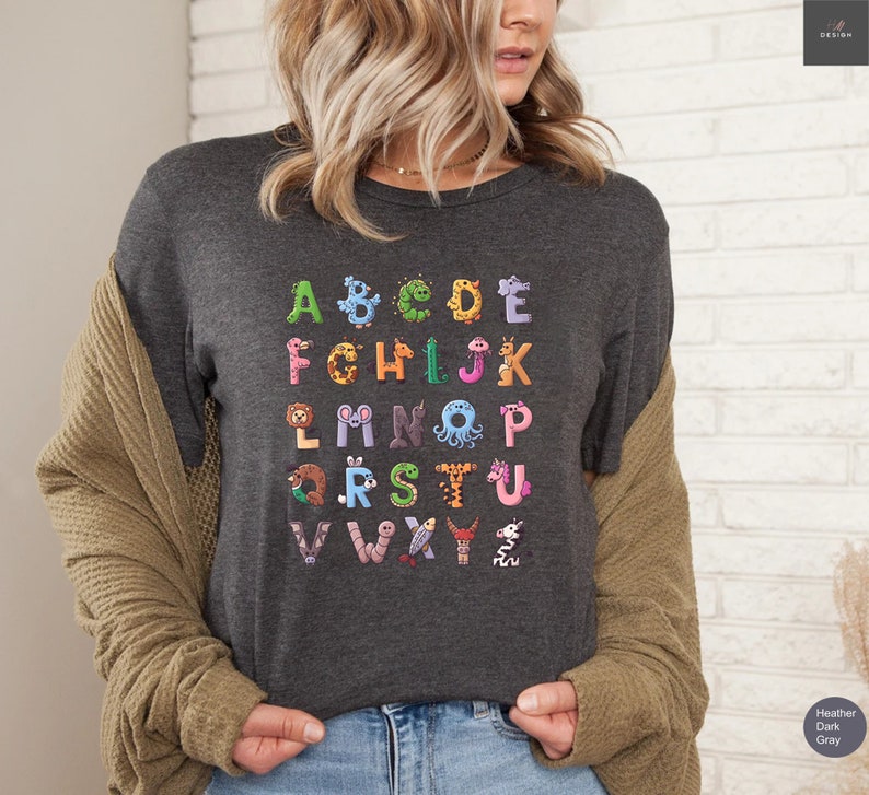 May include: Dark gray t-shirt featuring a colorful alphabet design. Each letter is creatively illustrated as an animal or creature, including a giraffe, octopus, and worm. The shirt is made of a soft material.