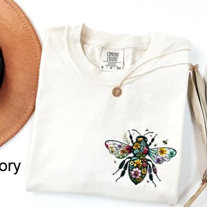 May include: Ivory-colored t-shirt featuring a colorful floral bee design. The shirt has a crew neck and short sleeves. A gold necklace with a small pendant is draped across the shirt. The words "COMFORT COLORS" are printed on a tag.
