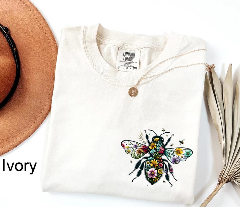 May include: Ivory-colored t-shirt featuring a colorful floral bee design. The shirt has a crew neck and short sleeves. A gold necklace with a small pendant is draped across the shirt. The words "COMFORT COLORS" are printed on a tag.
