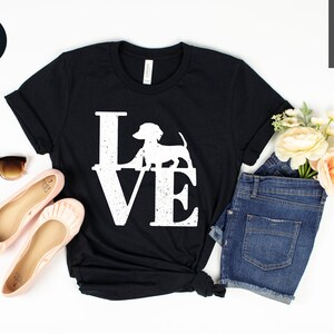 May include: Black t-shirt with the word "LOVE" in large white letters, featuring a dachshund silhouette in place of the "O". The shirt is styled with sunglasses, pink flats, and denim shorts. A black circle with the word "Black" is in the upper left corner.