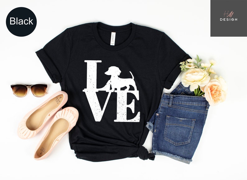 May include: Black t-shirt with the word "LOVE" in large white letters, featuring a dachshund silhouette in place of the "O". The shirt is styled with sunglasses, pink flats, and denim shorts. A black circle with the word "Black" is in the upper left corner.