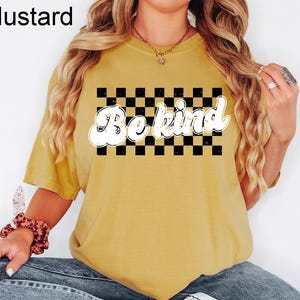 Pu&ograve; includere: Una t-shirt a maniche corte color giallo senape con una grafica rettangolare a scacchi bianchi e neri sul petto. Il testo "Be kind" &egrave; stampato in un carattere bianco, grassetto e in stile retr&ograve; sopra il centro del motivo.