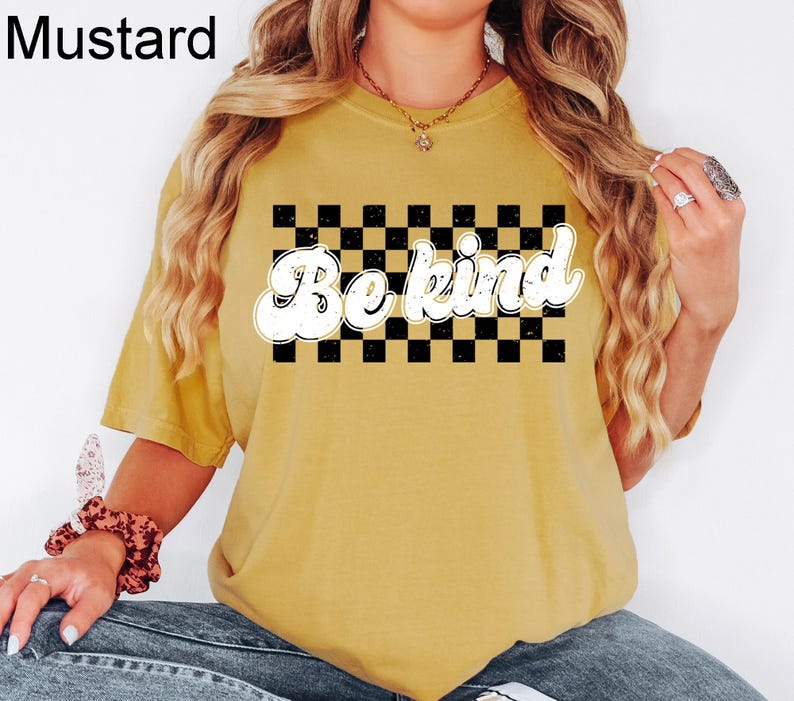 Pu&ograve; includere: Una t-shirt a maniche corte color giallo senape con una grafica rettangolare a scacchi bianchi e neri sul petto. Il testo "Be kind" &egrave; stampato in un carattere bianco, grassetto e in stile retr&ograve; sopra il centro del motivo.
