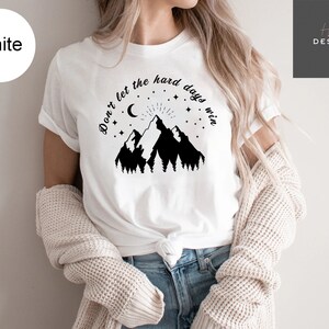 Pu&ograve; includere: T-shirt bianca con grafica di montagna nera, luna crescente, stelle e la scritta "Don't let the hard days win." La maglietta &egrave; realizzata in materiale morbido ed &egrave; adatta per l'abbigliamento casual.