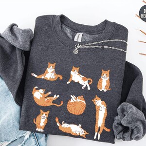 May include: A dark heather gray crewneck sweatshirt featuring a graphic print of seven orange and white tabby cats in various poses. The sweatshirt has a round neckline and long sleeves, with a small circular tag in the upper right corner labeled Dark Heather.