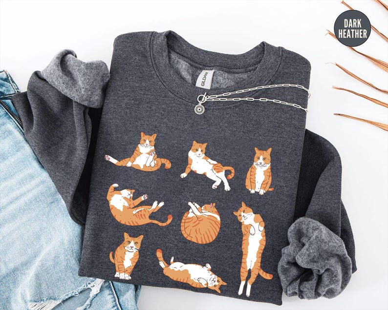 May include: A dark heather gray crewneck sweatshirt featuring a graphic print of seven orange and white tabby cats in various poses. The sweatshirt has a round neckline and long sleeves, with a small circular tag in the upper right corner labeled Dark Heather.