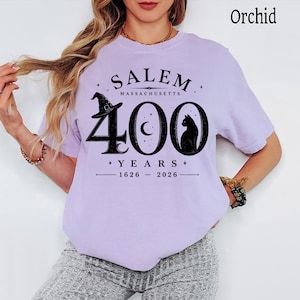 Puede incluir: Una camiseta color orqu&iacute;dea con un estampado gr&aacute;fico negro que dice SALEM MASSACHUSETTS 400 YEARS 1626-2026. El dise&ntilde;o incluye un sombrero de bruja, una luna creciente y la silueta de un gato integrados en el n&uacute;mero grande 400.