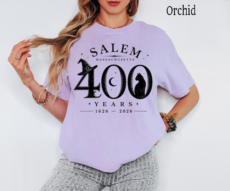 Puede incluir: Una camiseta color orqu&iacute;dea con un estampado gr&aacute;fico negro que dice SALEM MASSACHUSETTS 400 YEARS 1626-2026. El dise&ntilde;o incluye un sombrero de bruja, una luna creciente y la silueta de un gato integrados en el n&uacute;mero grande 400.