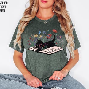May include: Heather forest green t-shirt featuring a black cat lying on an open book, surrounded by colorful flowers and butterflies. The text "Heather Forest Green" is printed on the top left corner of the image. A whimsical design for cat lovers.