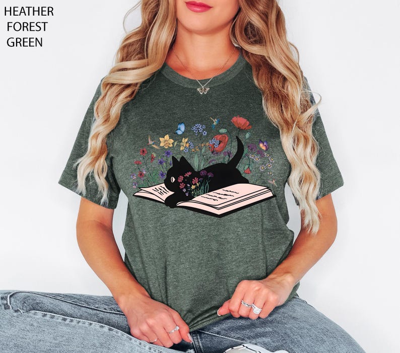 May include: Heather forest green t-shirt featuring a black cat lying on an open book, surrounded by colorful flowers and butterflies. The text "Heather Forest Green" is printed on the top left corner of the image. A whimsical design for cat lovers.