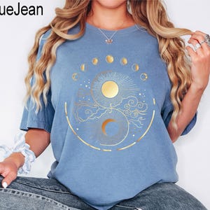 Pu&ograve; includere: T-shirt azzurra con un disegno dorato di sole e luna, circondato da fasi lunari e nuvole. Il testo "BlueJean" &egrave; nell'angolo in alto a sinistra.