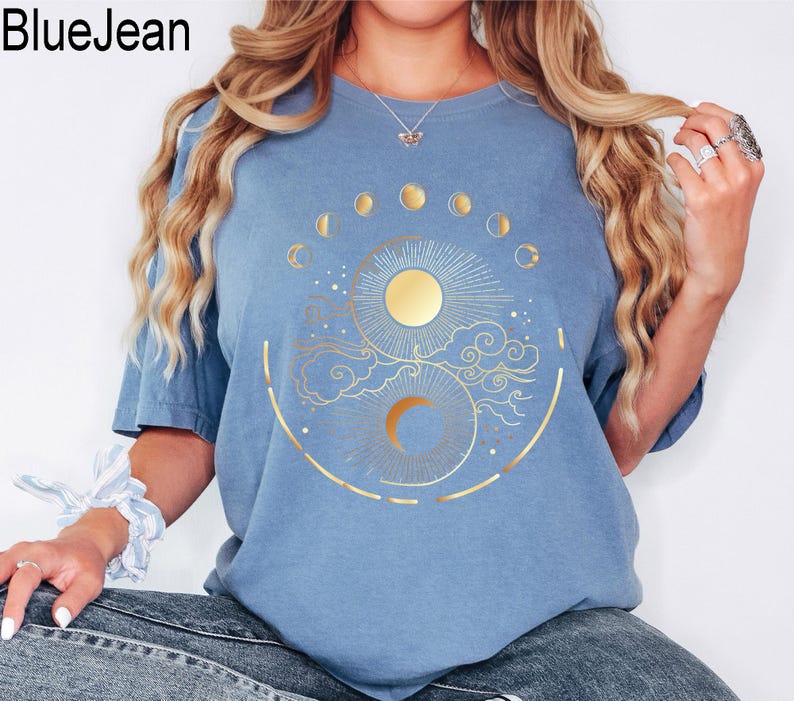 Pu&ograve; includere: T-shirt azzurra con un disegno dorato di sole e luna, circondato da fasi lunari e nuvole. Il testo "BlueJean" &egrave; nell'angolo in alto a sinistra.
