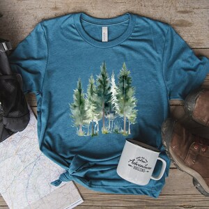May include: Teal-colored t-shirt featuring a watercolor illustration of several evergreen trees. The shirt is paired with a white enamel mug that reads "And so the adventure begins." Hiking boots and a backpack are also visible.